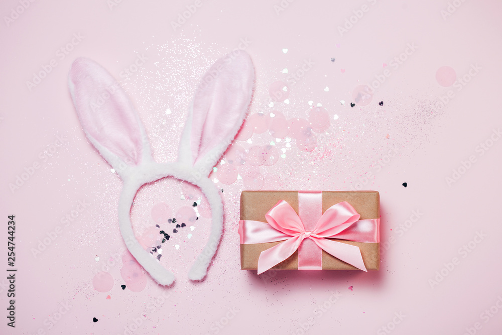 Top view and flat lay of Easter symbol - bunny ears and gift box on pink background. Festive and bright, confetti and sparks. Easter greetings Top view and flat lay of Easter symbol - bunny ears and gift box on pink background. Festive and bright, confetti and sparks. Easter greetings