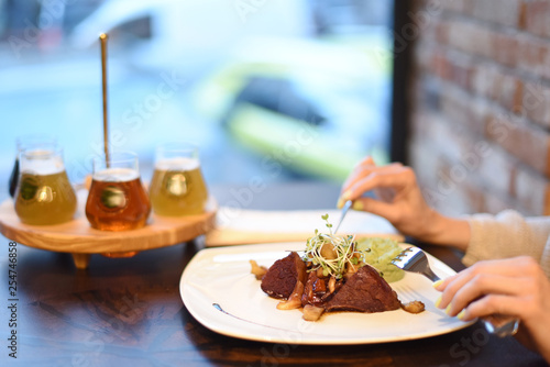 Meat steak on a dish in a restaurant. Girl's hands with a fork and knife. In the background a tasting beer set without focus.