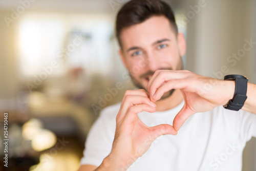 Young handsome man wearing casual white t-shirt at home smiling in love showing heart symbol and shape with hands. Romantic concept.