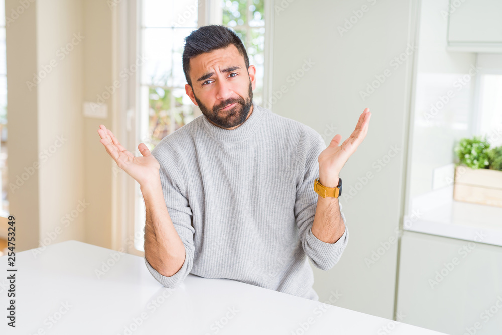 Handsome hispanic man wearing casual sweater at home clueless and confused expression with arms and hands raised. Doubt concept. Handsome hispanic man wearing casual sweater at home clueless and confused expression with arms and hands raised. Doubt concept.