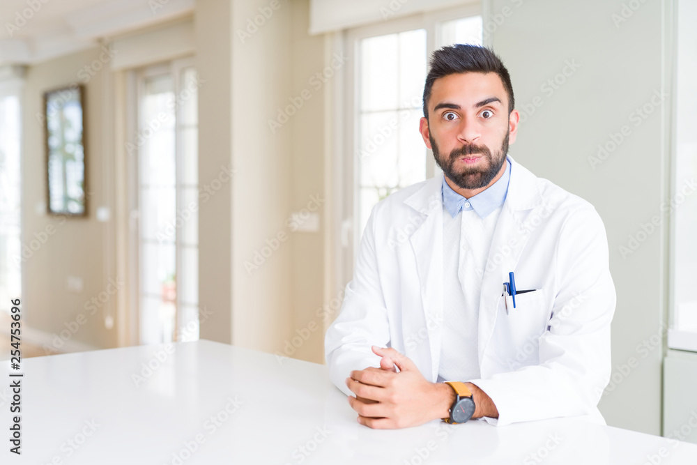 Handsome hispanic doctor or therapist man wearing medical coat at the clinic puffing cheeks with funny face. Mouth inflated with air, crazy expression. Handsome hispanic doctor or therapist man wearing medical coat at the clinic puffing cheeks with funny face. Mouth inflated with air, crazy expression.