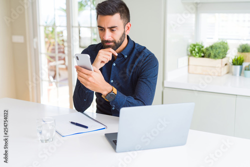 Handsome hispanic business man using smartphone and laptop at the office serious face thinking about question, very confused idea