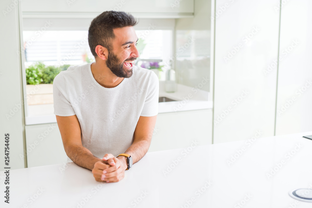 Handsome hispanic man casual white t-shirt at home looking away to side with smile on face, natural expression. Laughing confident.