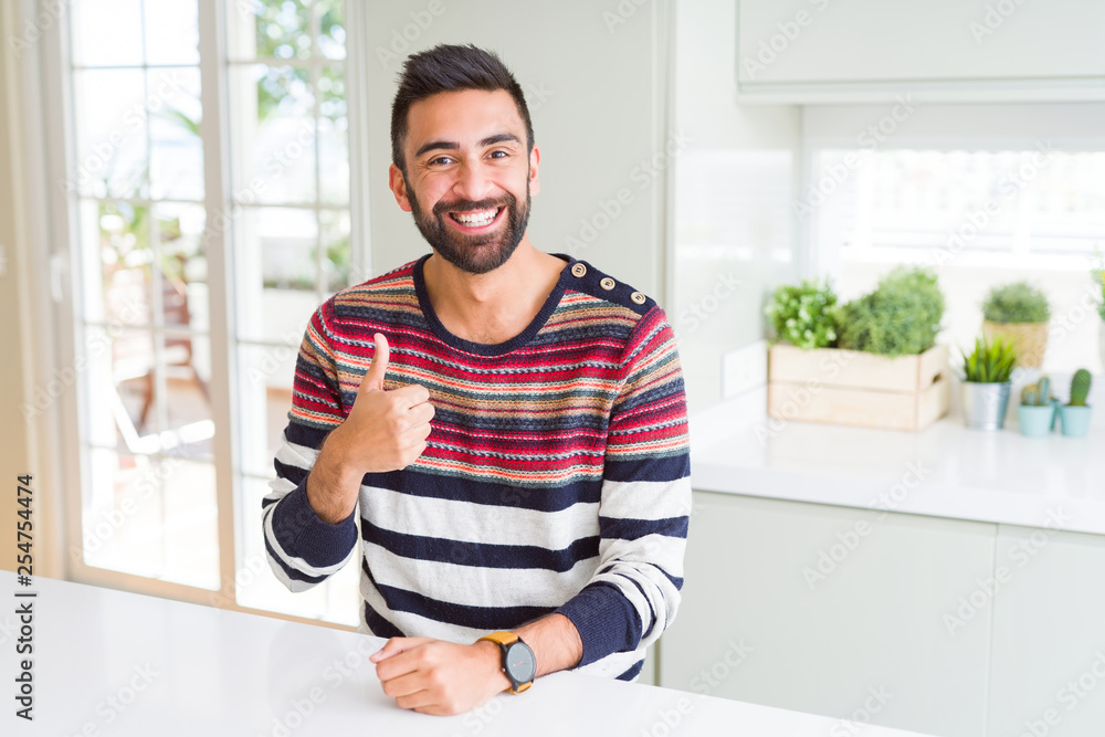 Handsome hispanic man wearing stripes sweater at home doing happy thumbs up gesture with hand. Approving expression looking at the camera with showing success. Handsome hispanic man wearing stripes sweater at home doing happy thumbs up gesture with hand. Approving expression looking at the camera with showing success.
