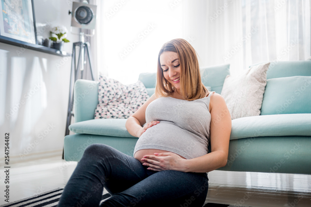 Beautiful smiling pregnant woman sitting on floor at home, touching and looking on belly Beautiful smiling pregnant woman sitting on floor at home, touching and looking on belly