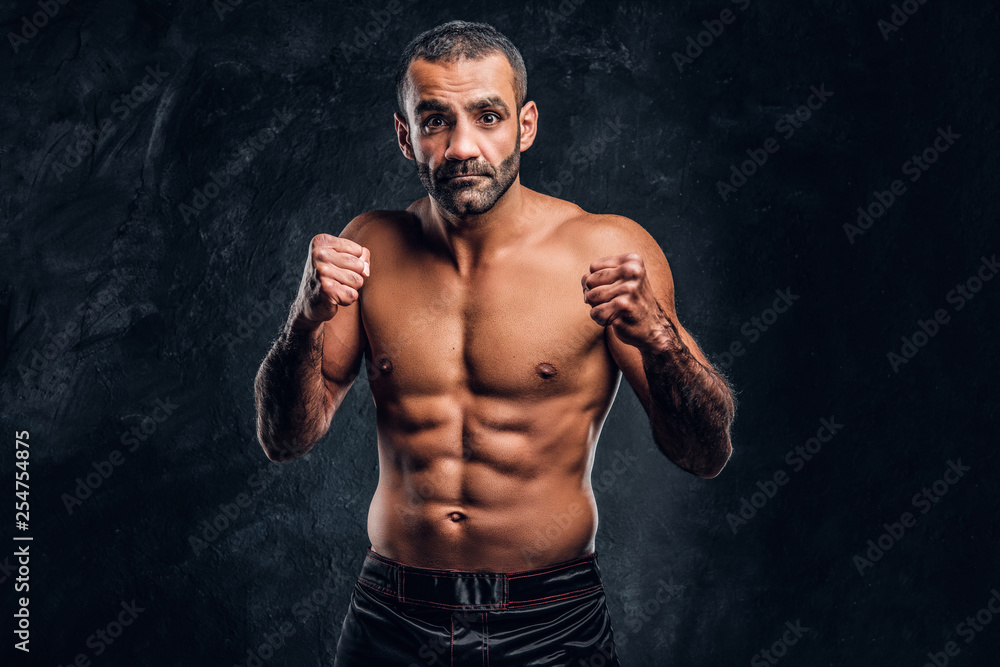Professional Muay Thai boxer with naked torso posing for a camera. Studio photo against a dark textured wall