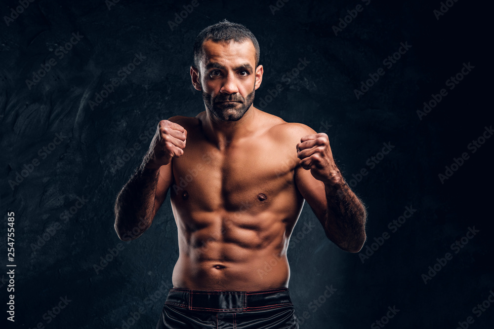 Portrait of a brutal professional fighter with naked torso posing for a camera. Studio photo against a dark textured wall