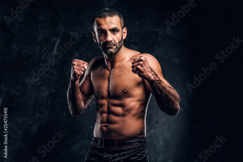 Professional Muay Thai boxer with naked torso posing for a camera. Studio photo against a dark textured wall © Fxquadro