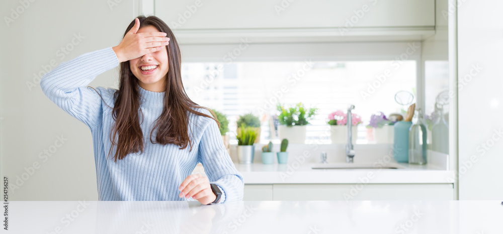 Wide angle picture of beautiful young woman sitting on white table at home smiling and laughing with hand on face covering eyes for surprise. Blind concept. Wide angle picture of beautiful young woman sitting on white table at home smiling and laughing with hand on face covering eyes for surprise. Blind concept.