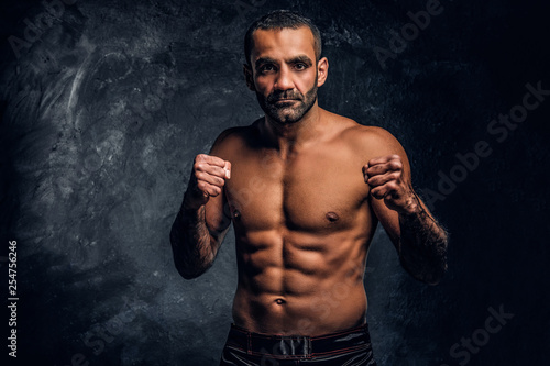 Professional Muay Thai boxer with naked torso posing for a camera. Studio photo against a dark textured wall © Fxquadro