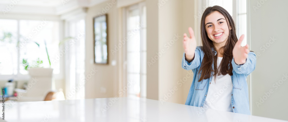 Wide angle picture of beautiful young woman sitting on white table at home looking at the camera smiling with open arms for hug. Cheerful expression embracing happiness. Wide angle picture of beautiful young woman sitting on white table at home looking at the camera smiling with open arms for hug. Cheerful expression embracing happiness.