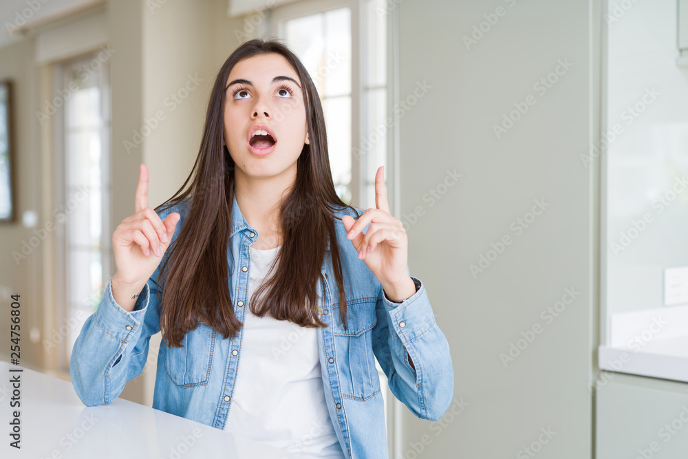 Beautiful young woman sitting on white table at home amazed and surprised looking up and pointing with fingers and raised arms. Beautiful young woman sitting on white table at home amazed and surprised looking up and pointing with fingers and raised arms.