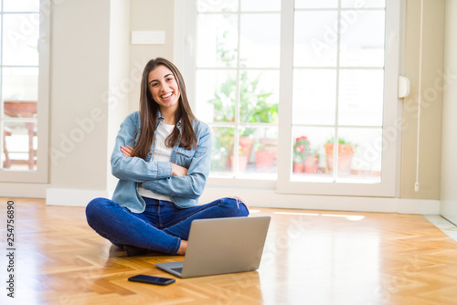 Beautiful young woman sitting on the floor with crossed legs using laptop happy face smiling with crossed arms looking at the camera. Positive person.