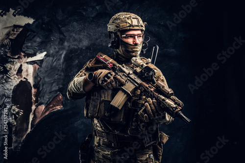 Fully equipped special forces soldier in camouflage uniform holding an assault rifle. Studio photo against a dark wall