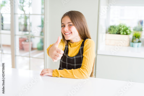 Beautiful young girl kid sitting on the table doing happy thumbs up gesture with hand. Approving expression looking at the camera showing success.