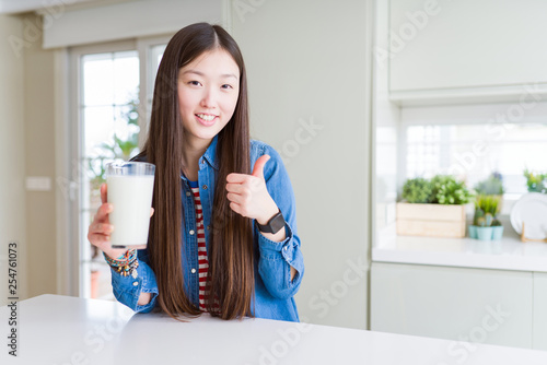 Beautiful Asian woman drinking a fresh glass of milk happy with big smile doing ok sign  thumb up with fingers  excellent sign