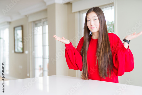 Beautiful Asian woman wearing red sweater on white table clueless and confused expression with arms and hands raised. Doubt concept.