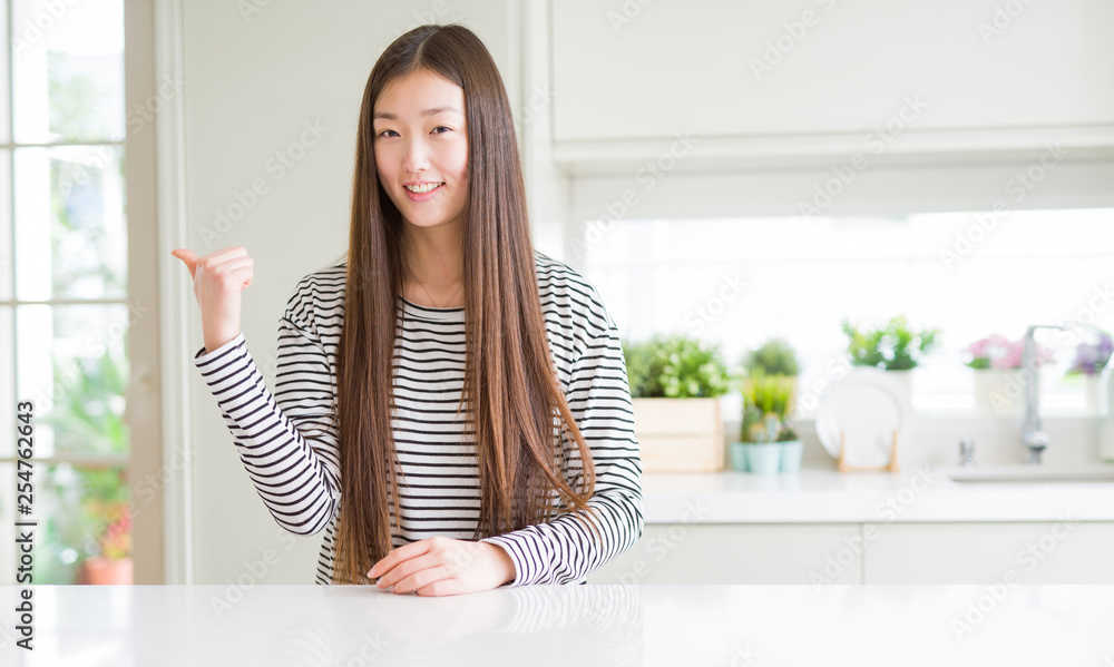 Beautiful Asian woman wearing stripes sweater smiling with happy face looking and pointing to the side with thumb up.