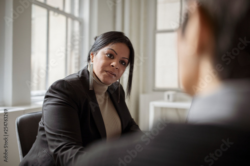 Female colleagues discussing while sitting against windows in office