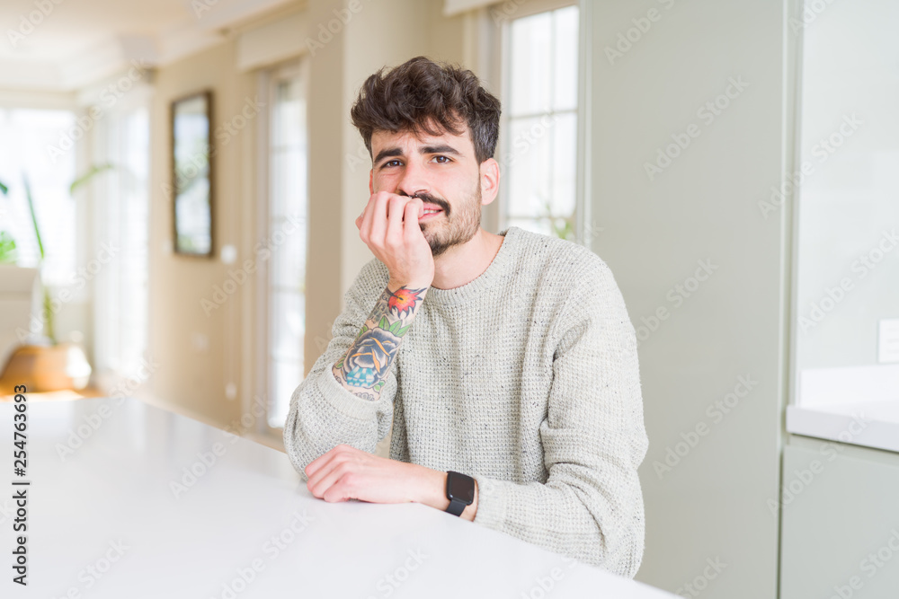 Young man wearing casual sweater sitting on white table looking stressed and nervous with hands on mouth biting nails. Anxiety problem. Young man wearing casual sweater sitting on white table looking stressed and nervous with hands on mouth biting nails. Anxiety problem.