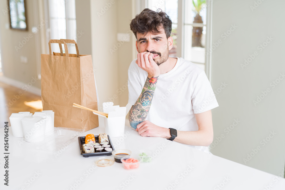 Young man eating asian sushi from home delivery looking stressed and nervous with hands on mouth biting nails. Anxiety problem. Young man eating asian sushi from home delivery looking stressed and nervous with hands on mouth biting nails. Anxiety problem.