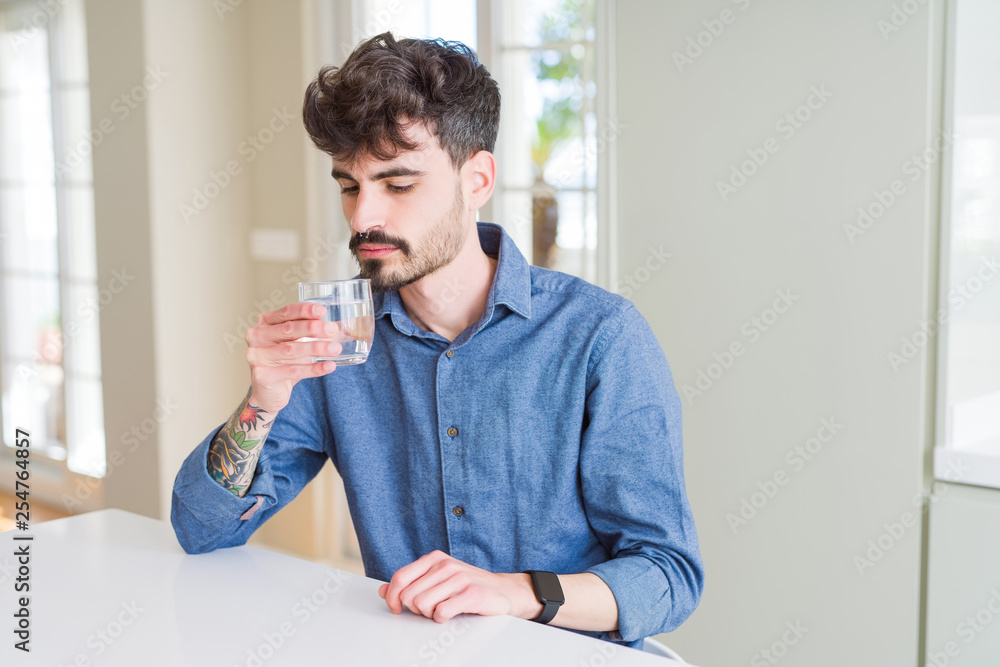 Young man drinking a fresh glass of water with a confident expression on smart face thinking serious
