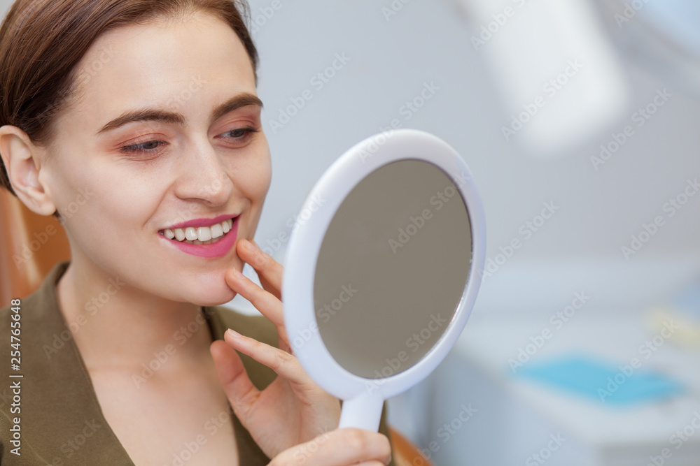Close up of a young beautiful woman smiling, looking in the mirror at the dental clinic. Attractive female patient examining her perfect whitened smile after medical treatment, Dental health concept Close up of a young beautiful woman smiling, looking in the mirror at the dental clinic. Attractive female patient examining her perfect whitened smile after medical treatment, Dental health concept