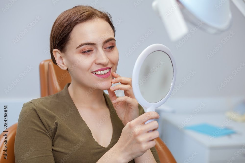 Attractive young woman smiling, examining her teeth in the mirror, while visiting dentist. Woman enjoying result of professional teeth cleaning and whitening, copy space. Health, wellness concept Attractive young woman smiling, examining her teeth in the mirror, while visiting dentist. Woman enjoying result of professional teeth cleaning and whitening, copy space. Health, wellness concept