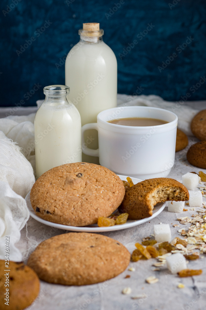 Cup of coffee with milk in a white cup, Bottles with milk, oatmeal cookies, oatmeal, raisins on a light background. Scene from breakfast Cup of coffee with milk in a white cup, Bottles with milk, oatmeal cookies, oatmeal, raisins on a light background. Scene from breakfast