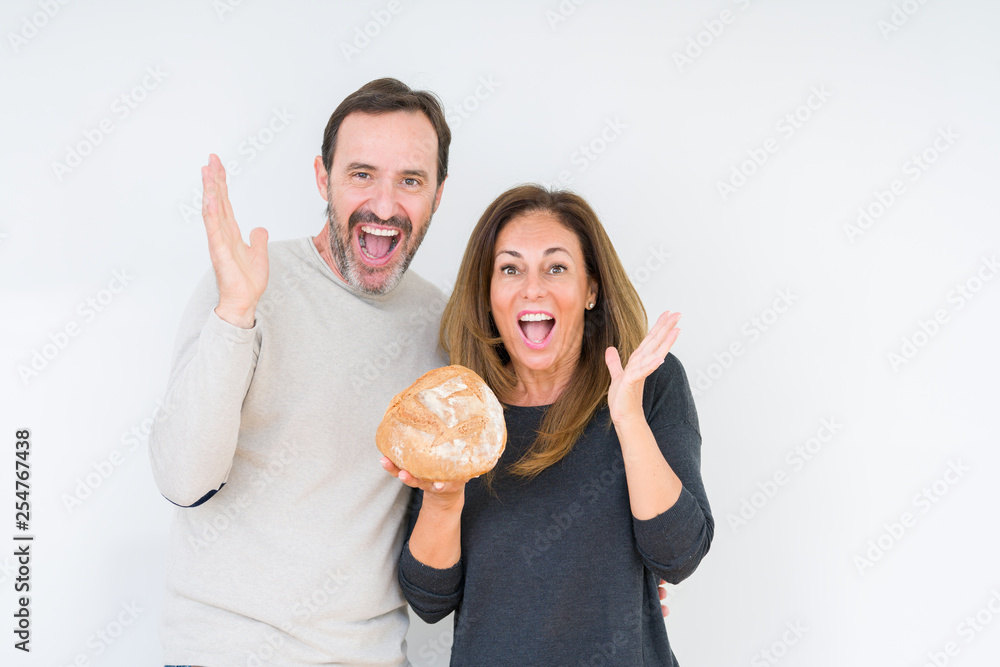 Middle age couple holding homemade fresh bread over isolated background very happy and excited, winner expression celebrating victory screaming with big smile and raised hands Middle age couple holding homemade fresh bread over isolated background very happy and excited, winner expression celebrating victory screaming with big smile and raised hands