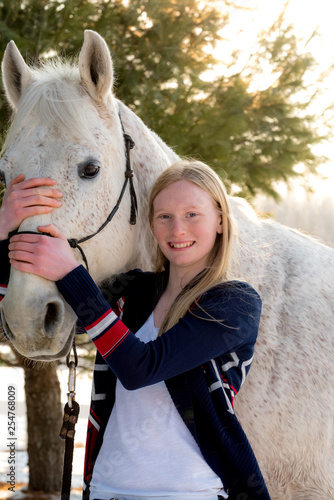 happy blonde teenager girl holding white/grey handsome horse. Sun is back lighting the scene and both are looking toward camera. 
