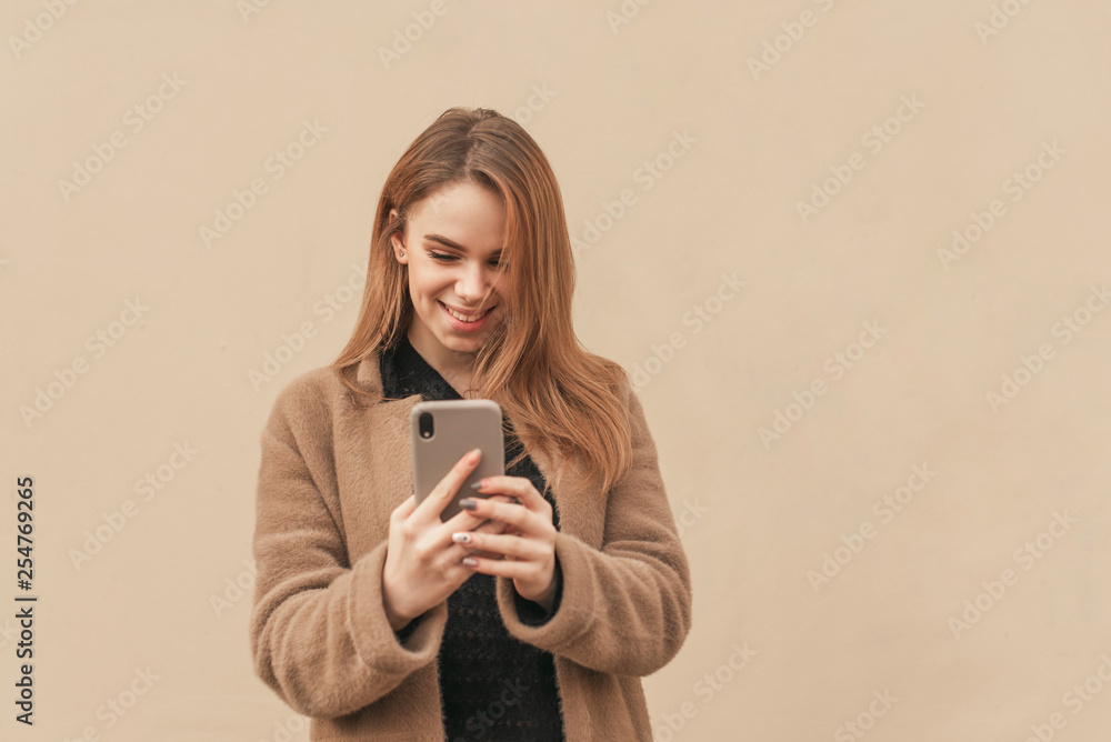 Happy girl in stylish clothes, wearing a coat,using smartphone on a beige background, looking at the screen and smiling. Lady in warm clothes and with a phone in hands, background of the beige wall Happy girl in stylish clothes, wearing a coat,using smartphone on a beige background, looking at the screen and smiling. Lady in warm clothes and with a phone in hands, background of the beige wall