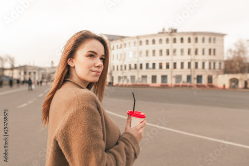 Smiling girl with a cup of coffee in his hand poses on the fnu of the street in a warm spring day, looking sideways. Attractive girl wears a coat, stands with a cup of coffee in her hands photo