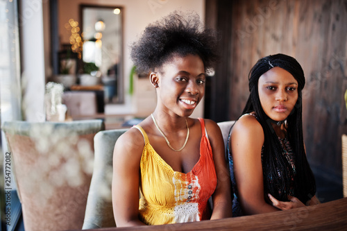 Two black african girlfriends at summer dresses sitting at table in cafe.