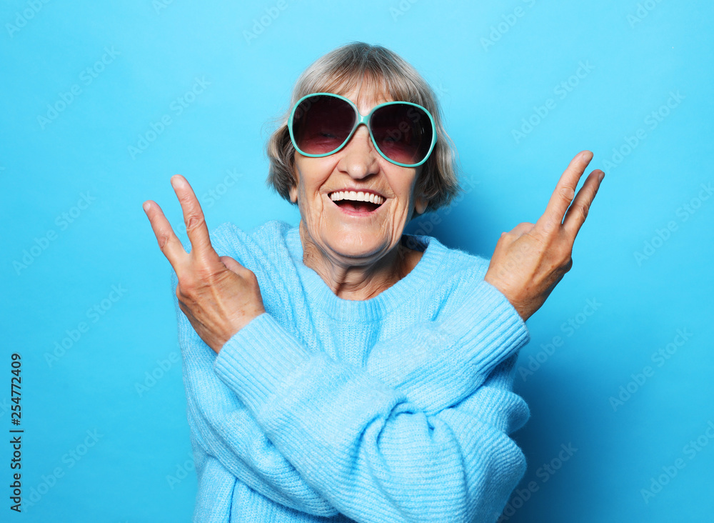 Funny old lady wearing blue sweater, hat and sunglasses showing victory sign. Isolated on blue background. Funny old lady wearing blue sweater, hat and sunglasses showing victory sign. Isolated on blue background.