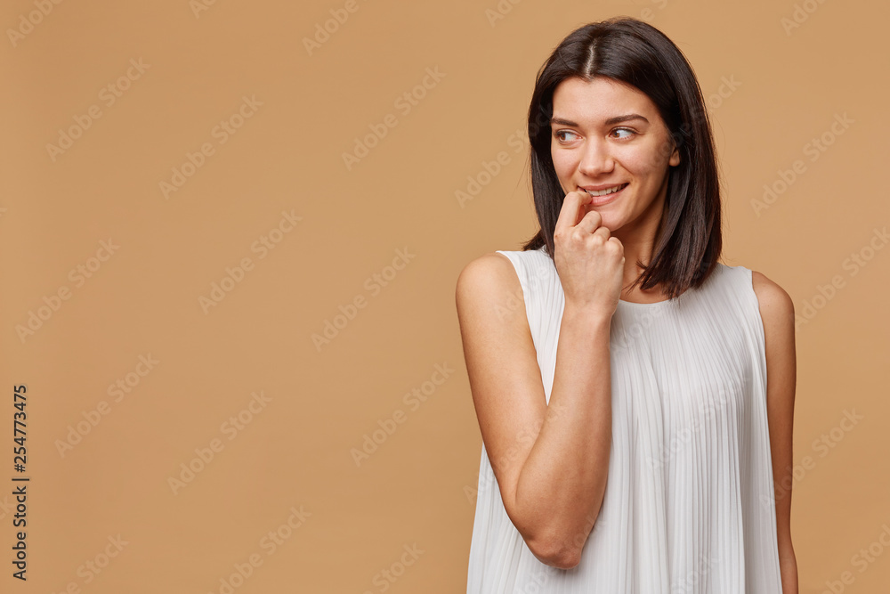 Happy excited thinking of something pleasant tanned woman standing holding a finger in her mouth, with warm smile looks to the left side of the empty space, over beige background Happy excited thinking of something pleasant tanned woman standing holding a finger in her mouth, with warm smile looks to the left side of the empty space, over beige background