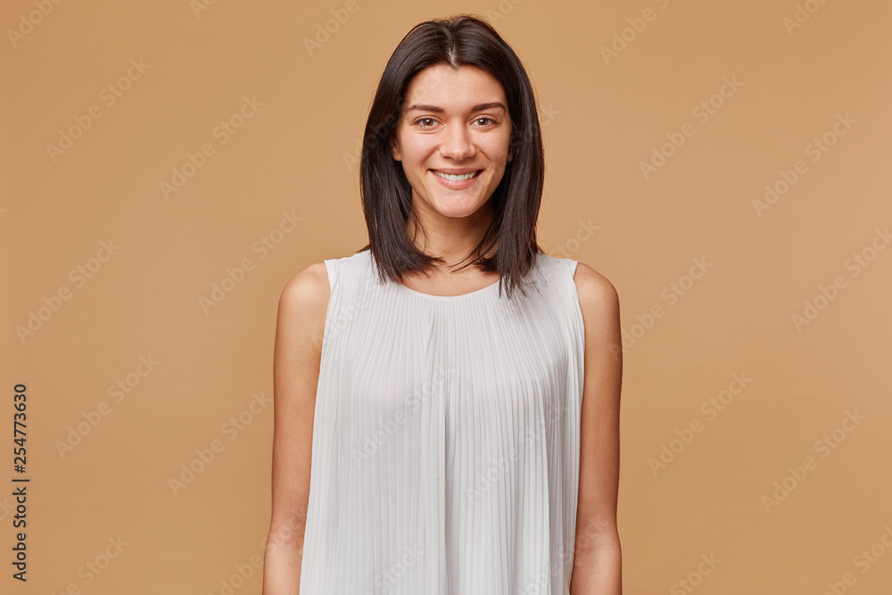 Portrait of a nice attractive beautiful girl with dark hair in white dress pleasantly smiling, isolated over beige background Portrait of a nice attractive beautiful girl with dark hair in white dress pleasantly smiling, isolated over beige background
