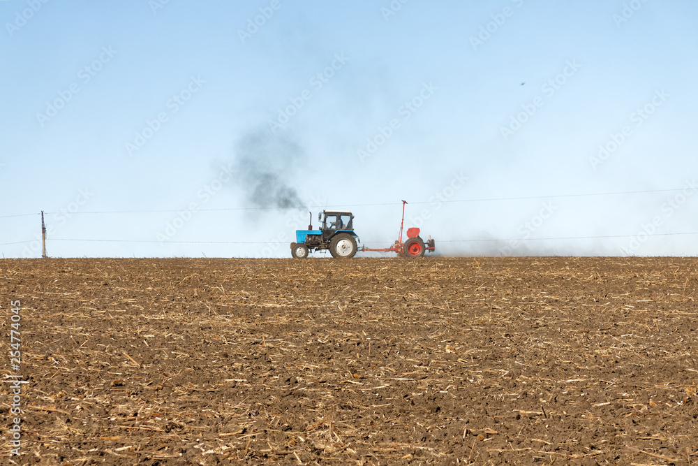 Fototapeta premium Blue tractor with a red seeder works on the field on a bright sunny autumn day