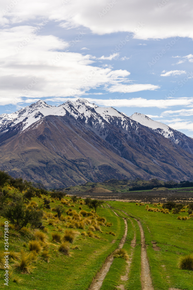 Vast valley with mountain range on the horizon, shadow play on mountain hills and dramatic sky during warm and sunny day, peaks covered with snow, road leading into mountains