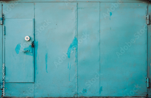 Closed imperfect blue metallic garage gate close-up. Texture of locked iron door. Cyan paint on grungy metal surface. Paint stains. Textured background of rough faded uneven surface of steel gates.