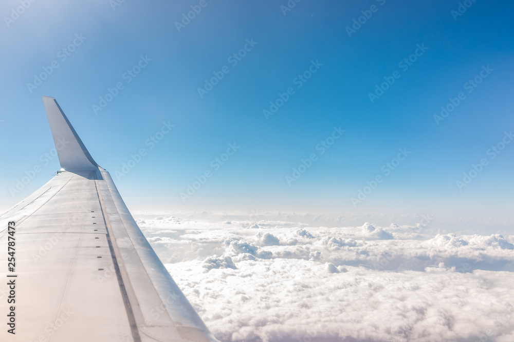 White blue airplane in sky with view from window high angle during sunny day with plane wing and clouds covering horizon