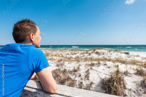 Destin Miramar beach city town village in Florida panhandle gulf of mexico ocean with back of young man closeup in blue shirt leaning on fence railing by sand dunes © Kristina Blokhin