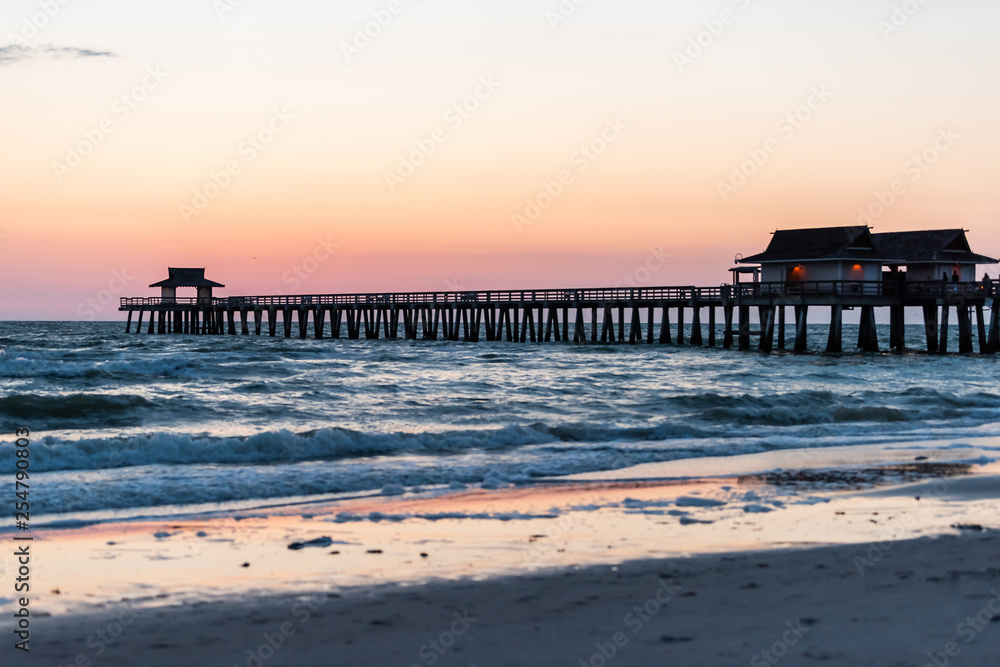 Naples, Florida pink and yellow pastel sunset twilight in gulf of Mexico with Pier and wooden pillars over horizon with dark blue ocean waves and tide