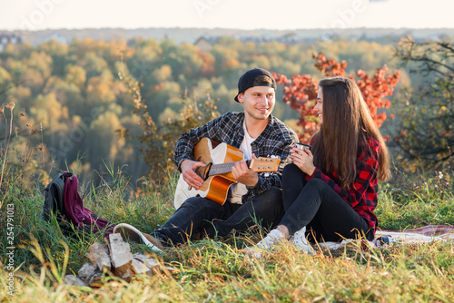 Young man playing on the guitar for his lovely girlfriend. Happy couple with guitar resting on picnic in spring park.