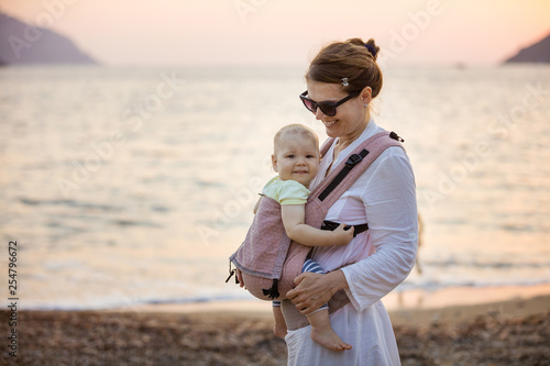 Photos Cheerful Caucasian woman with little daughter in baby carrier on beach at sunset