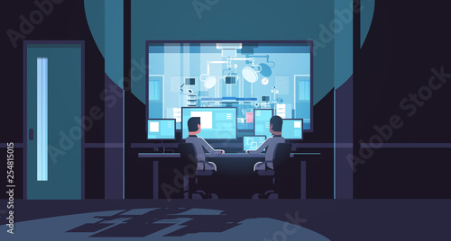 two men looking at monitors sitting behind glass window hospital operating table medical surgery room dark office interior surveillance security system flat horizontal