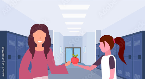schoolgirl giving apple to woman teacher in school lobby corridor interior with rows of blue lockers education concept horizontal portrait flat