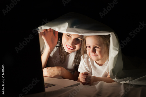 Happy mom and daughter watching a cartoon under the covers in bed