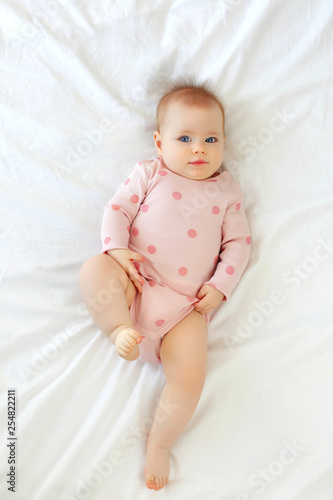 A charming blue-eyed baby with a funny hairstyle 9 months lies in bed in a pink body © dinaphoto