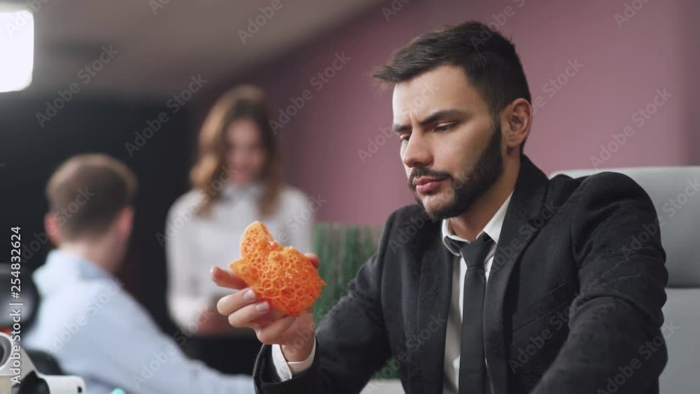 Young man designer in modern office holding in hands a plastic human heart model printed on a 3D printer, office workers on blurred background. Young man designer in modern office holding in hands a plastic human heart model printed on a 3D printer, office workers on blurred background.